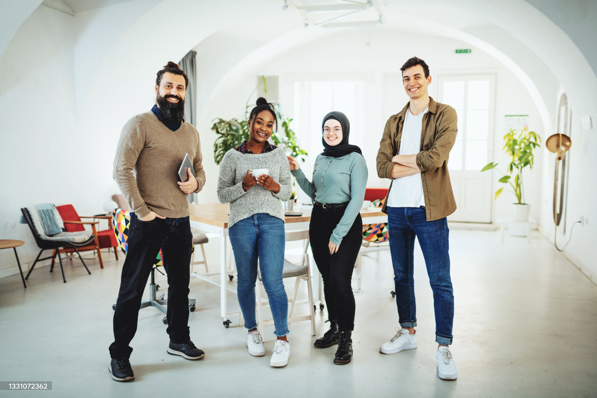 Four smiling professionals standing together in a bright, modern open-plan office with arched ceilings and collaborative workspace in the background.
